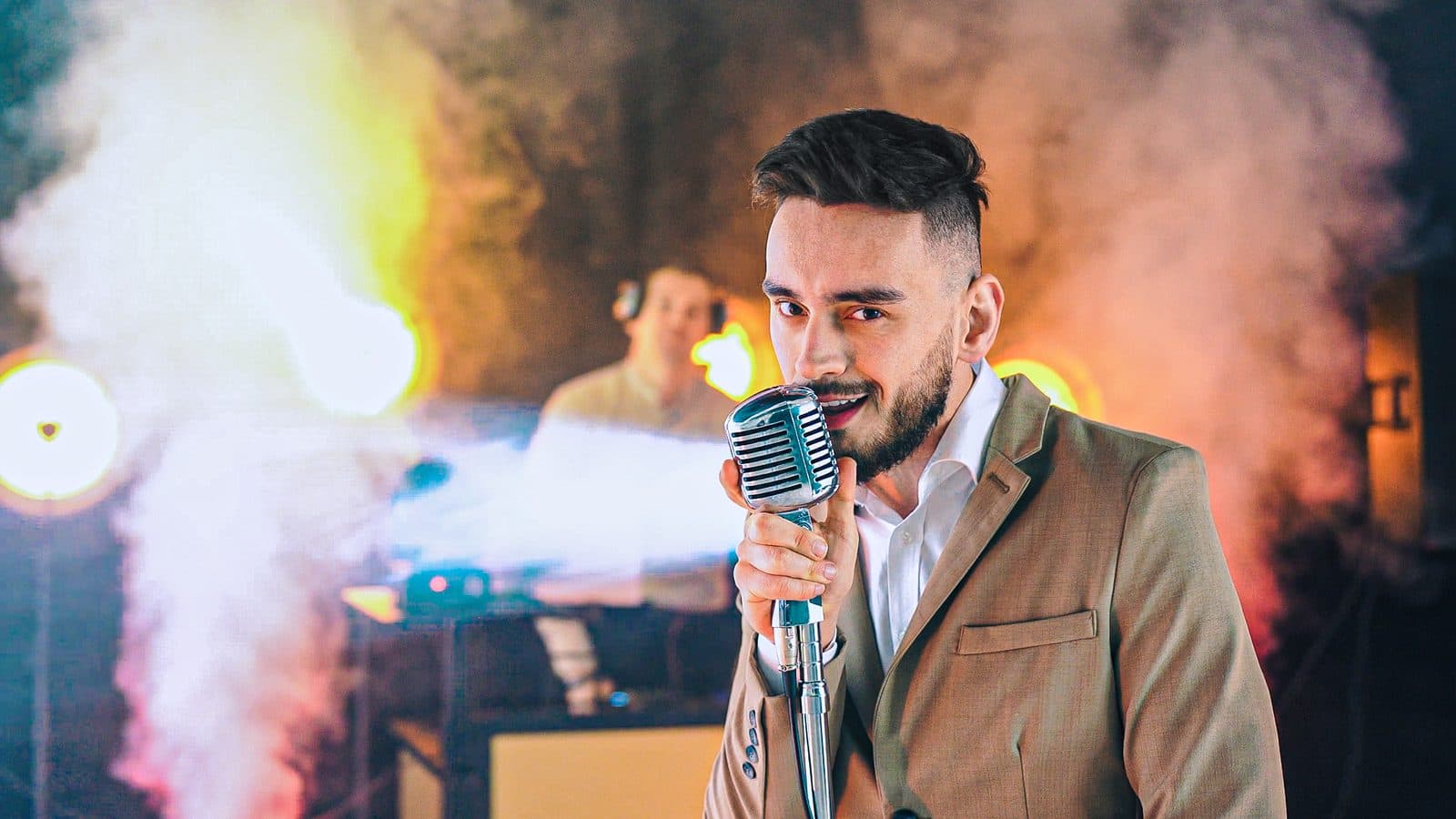 Denis Impulsywni śpiewa do zabytkowego mikrofonu na scenie w otoczeniu dymu i kolorowych świateł